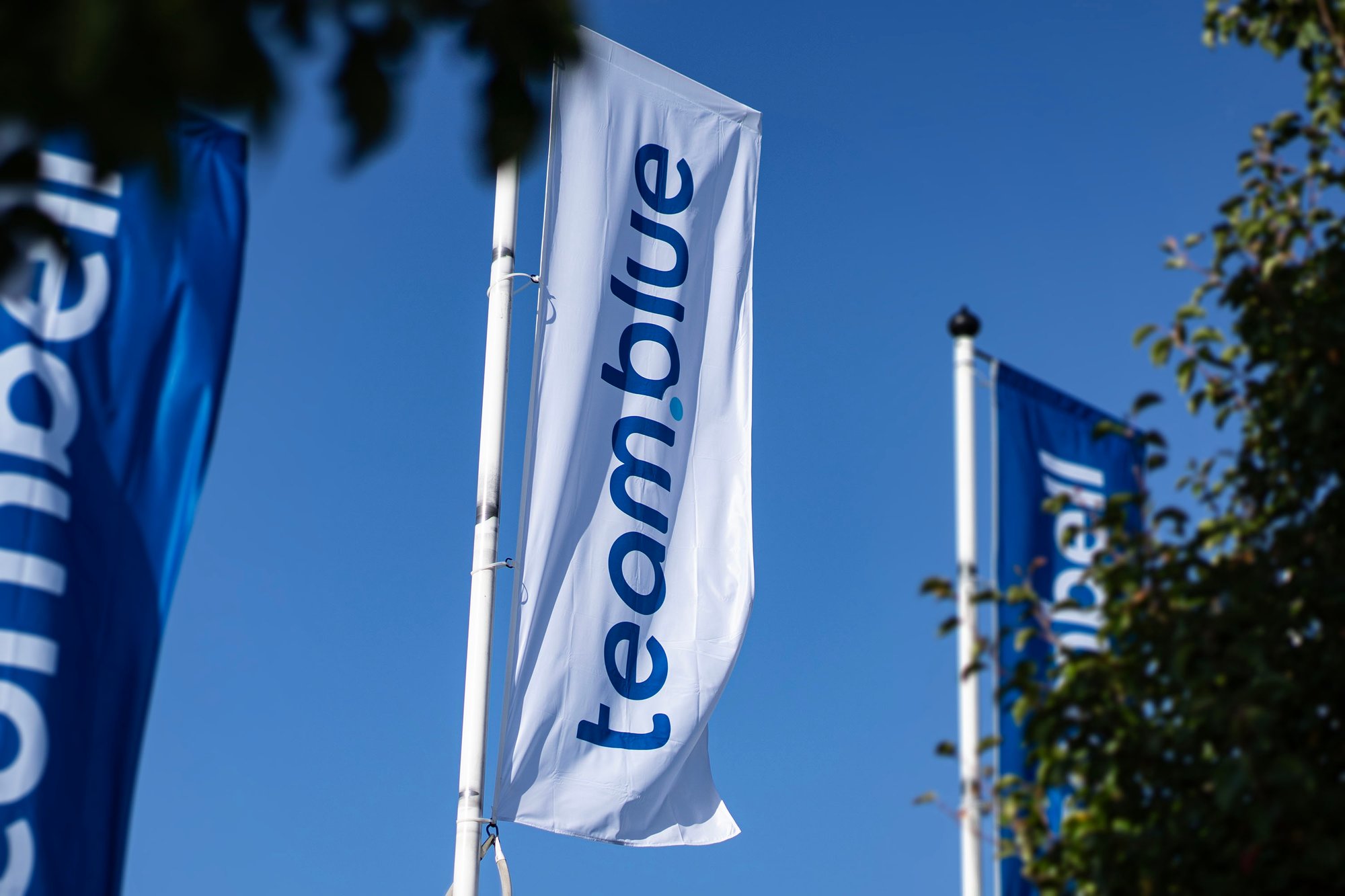 A white team.blue flag and a blue team.blue flag waving on flagpoles against a clear blue sky.